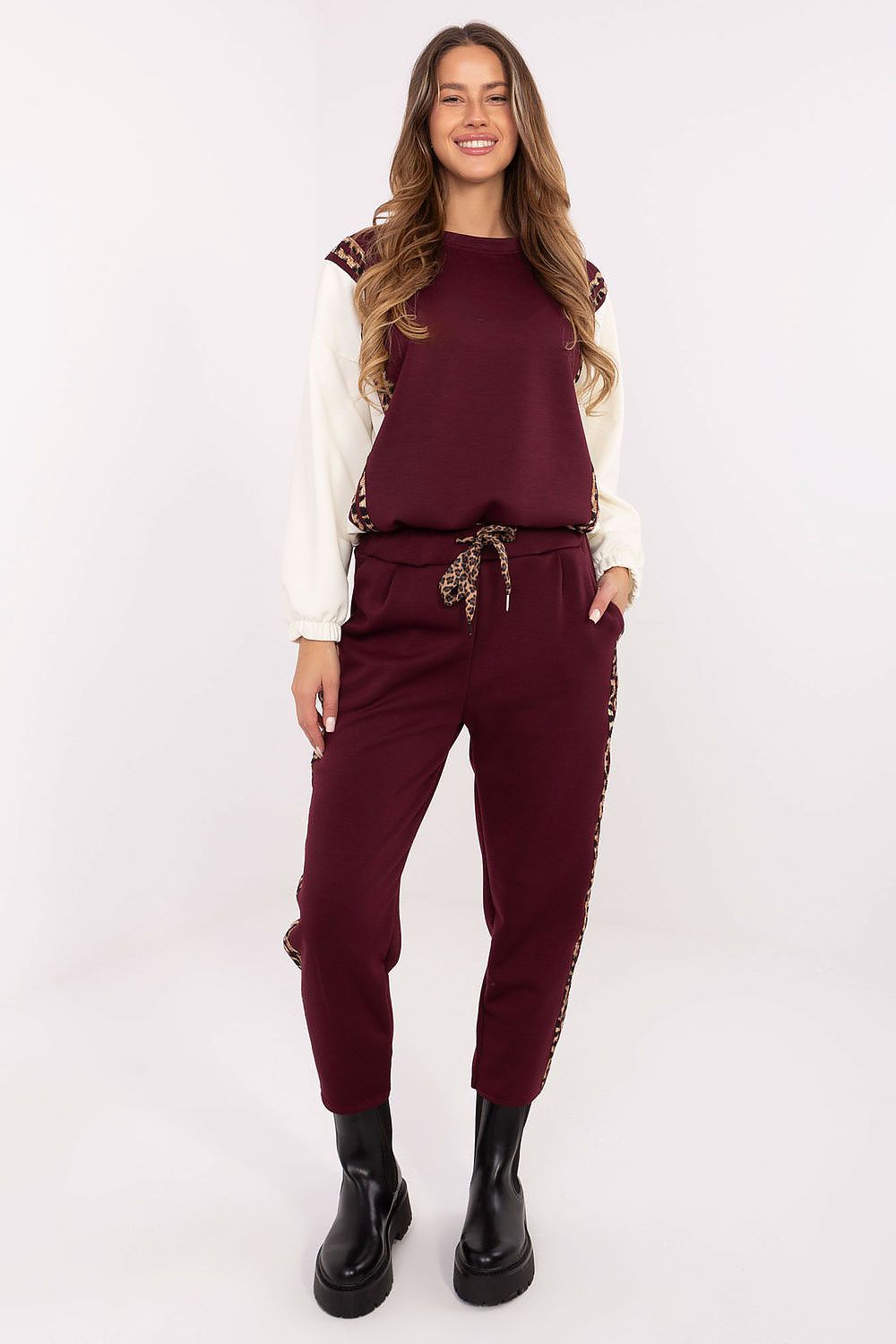 A woman modeling a burgundy two-piece lounge set consisting of a sleeveless top with white sleeves and matching jogger pants with gold chain accents, paired with black boots, against a plain white studio background.