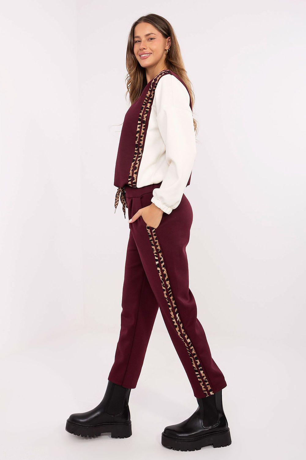 A woman modeling a matching burgundy two-piece set consisting of a sleeveless vest and jogger pants, both featuring a gold chain stripe down the side, styled with a white long-sleeve shirt and black platform boots against a white studio background.