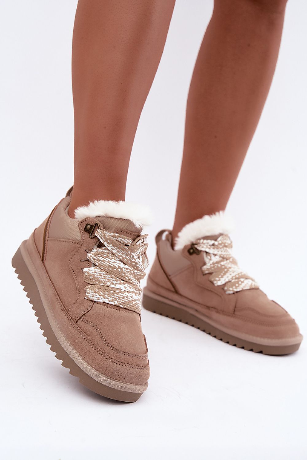 A pair of beige suede ankle boots with white faux-fur trim and chunky metallic silver laces, shown on a model's feet against a white studio background.
