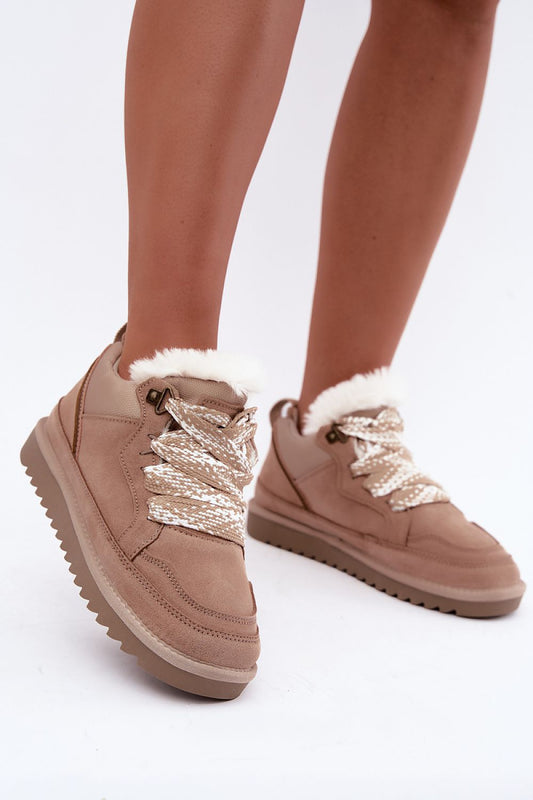 A pair of beige suede ankle boots with white faux-fur trim and chunky metallic silver laces, shown on a model's feet against a white studio background.