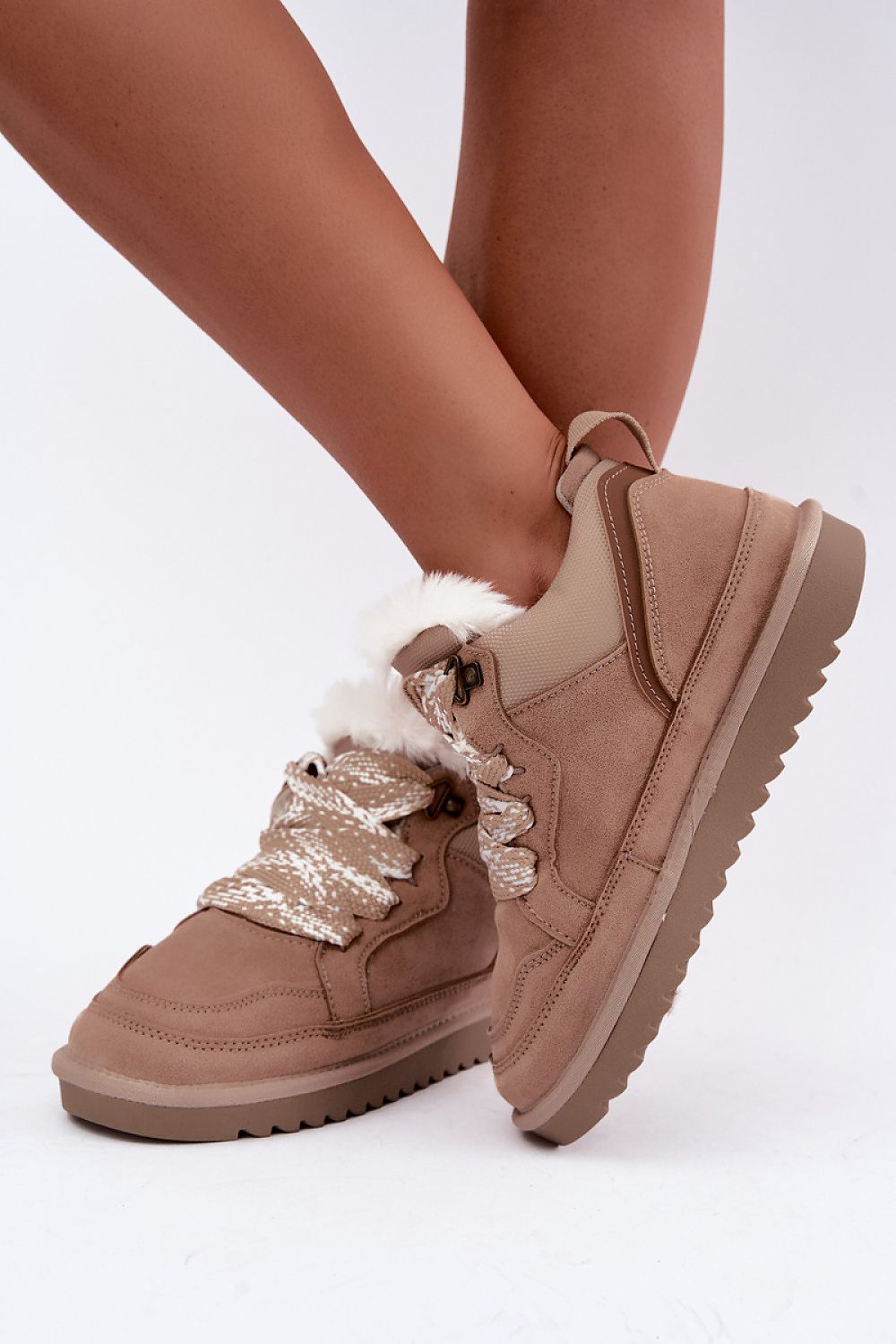 A pair of women's low-top winter sneakers in taupe brown suede, featuring a fluffy white faux-fur collar, chunky lug soles, and metallic gold patterned laces, shown on a model's feet against a white studio background.