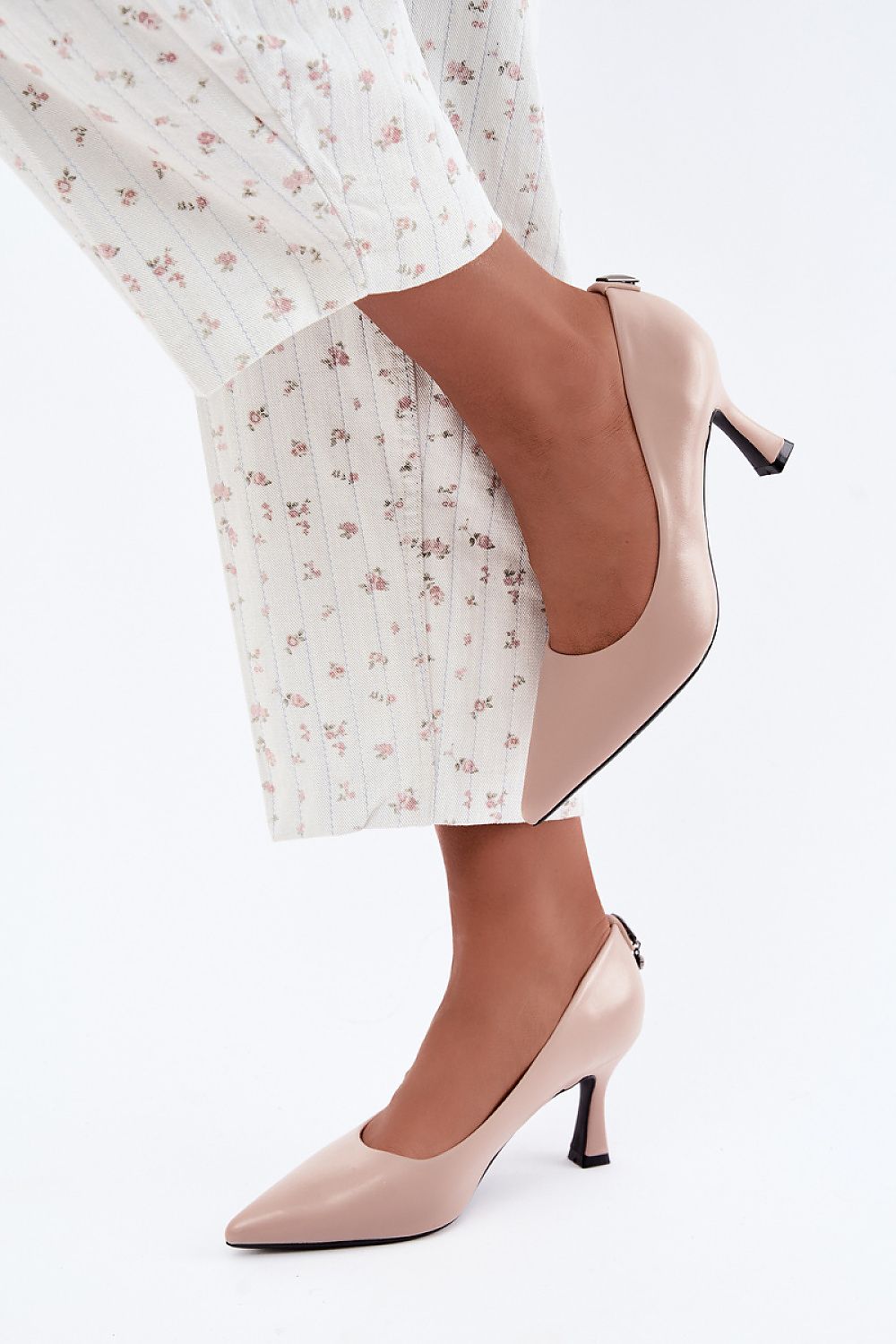 A person wearing light blush pink pointed-toe kitten heel pumps with a small buckle detail at the ankle, paired with white floral wide-leg trousers, against a clean white background.