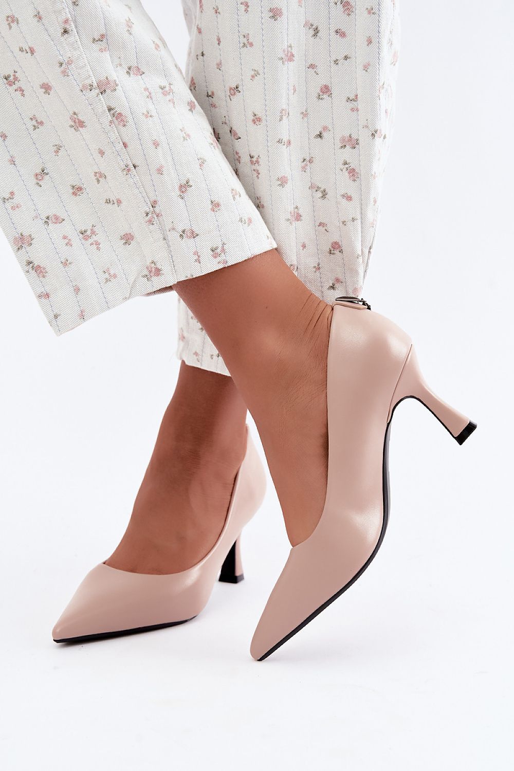 A close-up of a person wearing light blush pink pointed-toe pumps with a kitten heel, paired with white floral cropped trousers, against a clean white background.