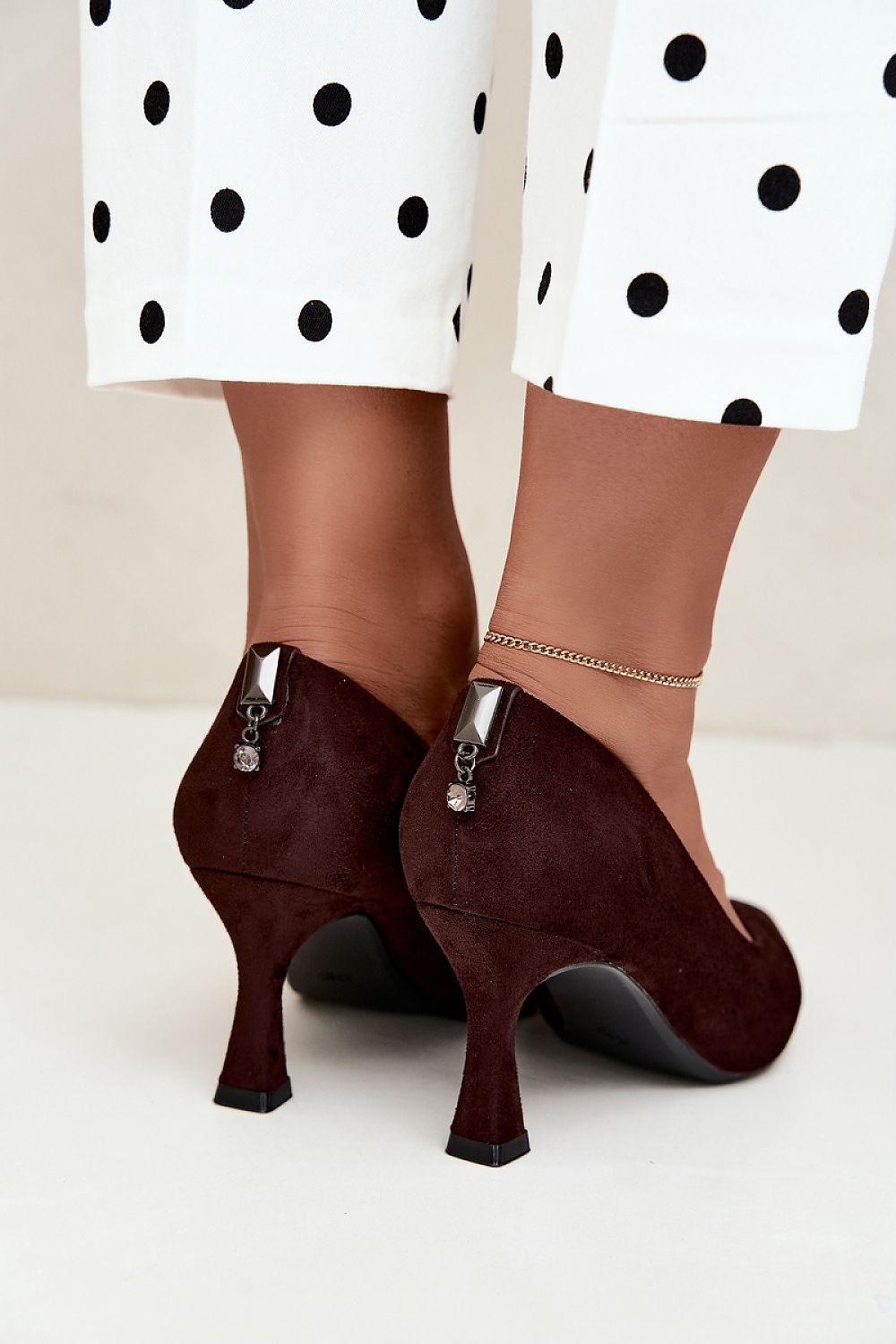 A close-up of a person's feet wearing dark burgundy suede high-heeled pumps with silver zipper details, styled with white and black polka dot trousers.