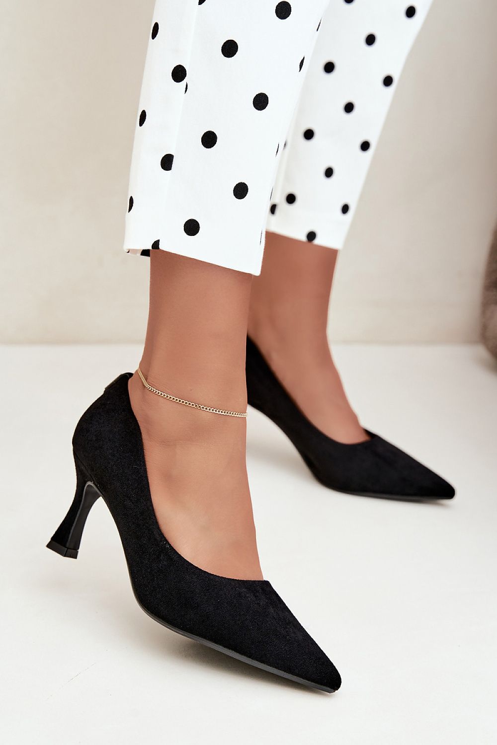 Black pointed-toe high heels with a kitten heel, shown on a model wearing white pants with black polka dots and a delicate silver anklet.