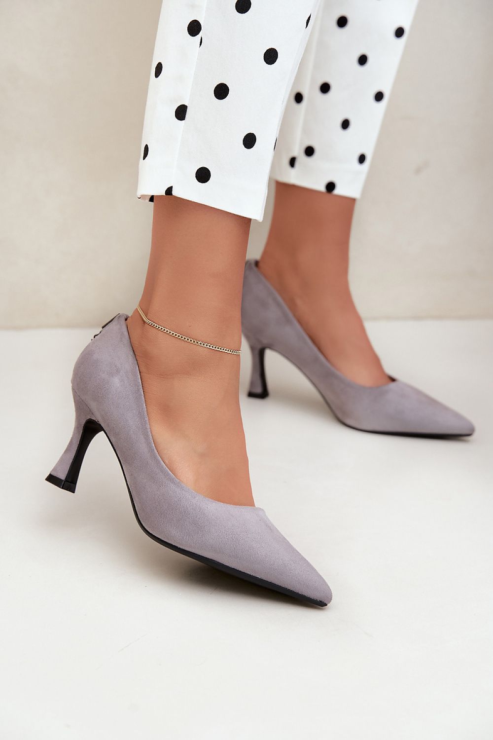 A person wearing light gray pointed-toe high heels with a kitten heel and a delicate ankle chain, paired with white pants featuring black polka dots, against a neutral beige background.