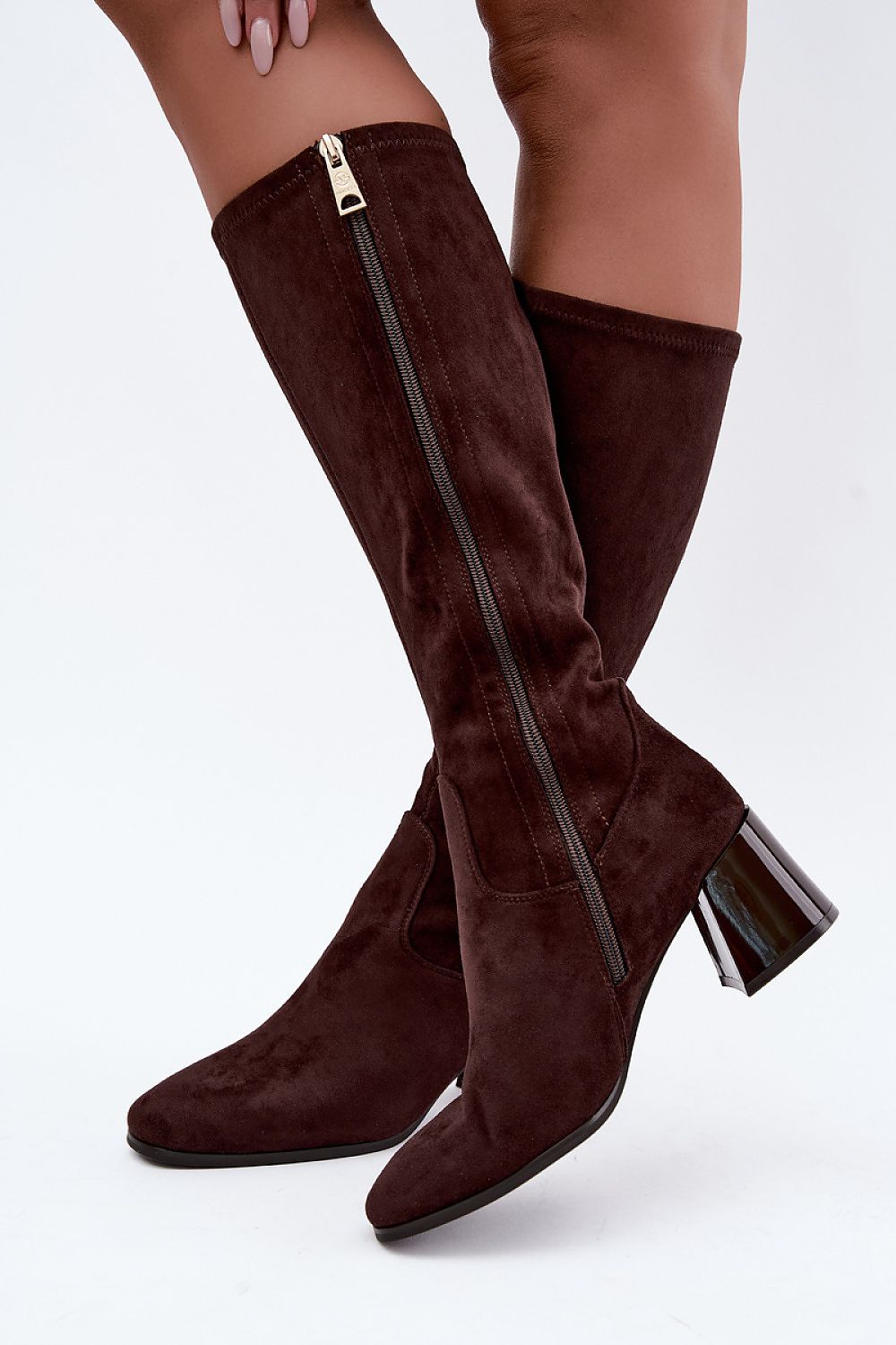 A pair of dark brown suede knee-high boots with side zippers and chunky block heels, shown on a model's legs against a white studio background.