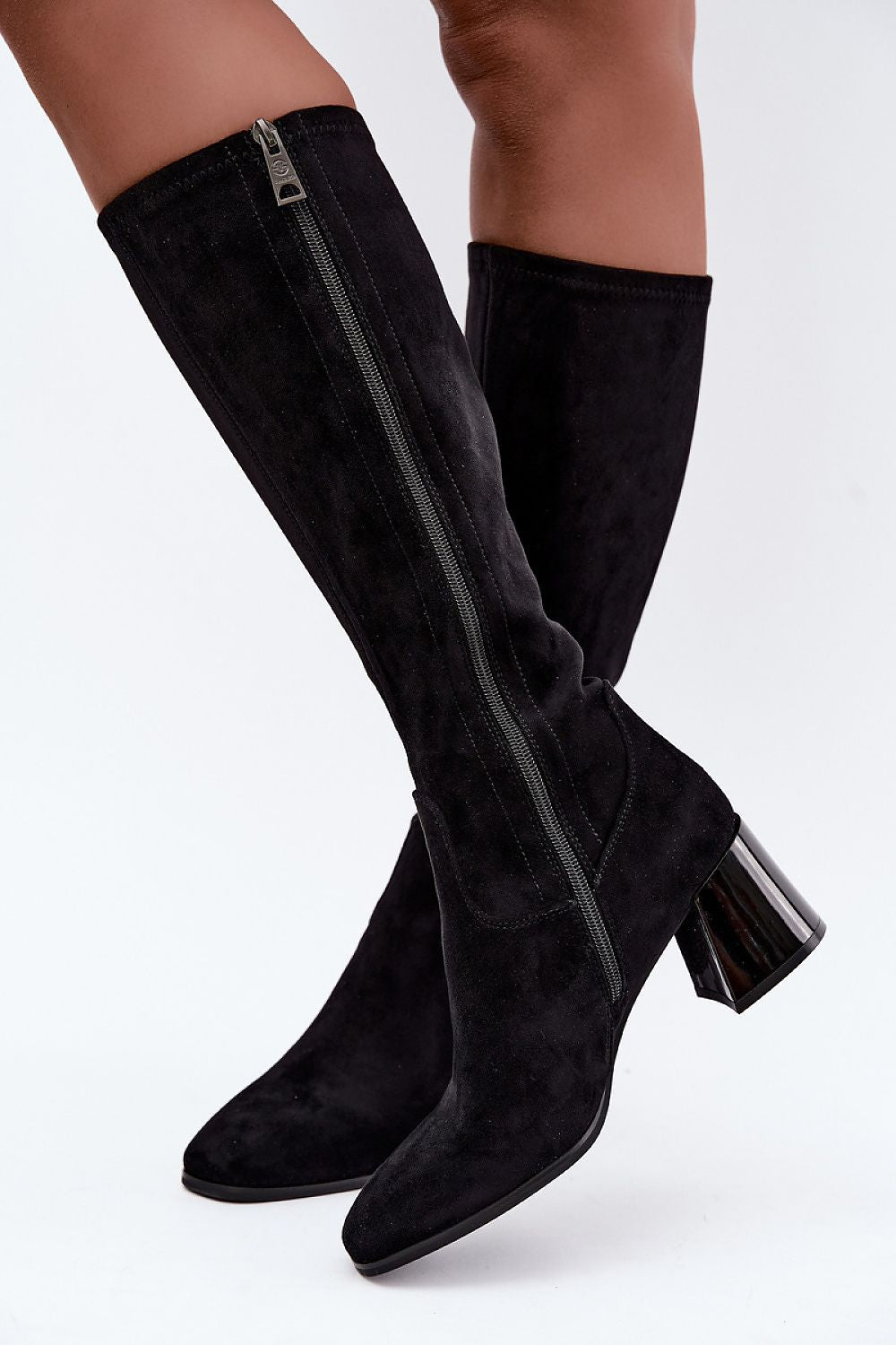Black suede knee-high boots with side zippers and chunky block heels, shown on a model's legs against a white studio background.