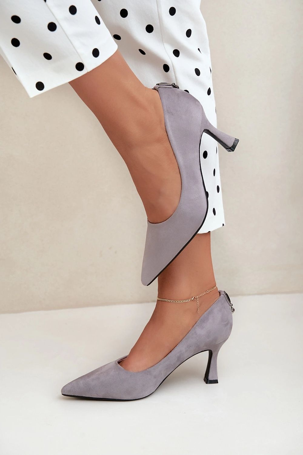 A person wearing light gray suede pointed-toe pumps with kitten heels, paired with white pants featuring black polka dots, against a neutral beige background.