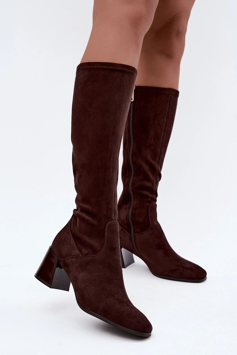 A pair of dark brown suede knee-high boots with block heels, shown on a model's legs against a plain white studio background.