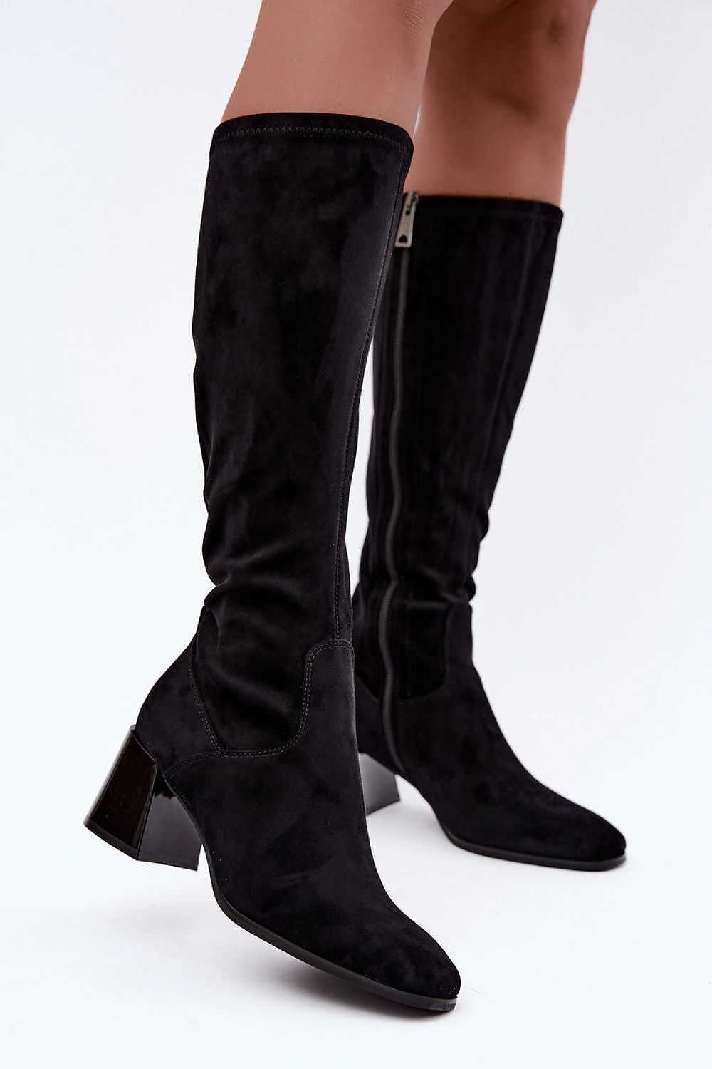 A pair of black suede knee-high boots with block heels, shown on a model's legs against a plain white studio background.