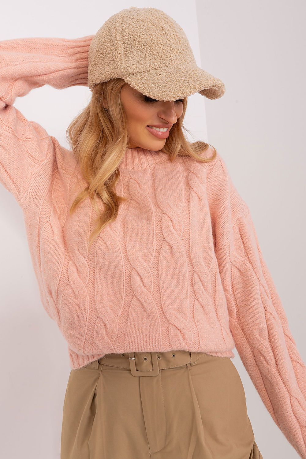 A woman wearing a dusty pink cable-knit sweater, a beige bouclé cap, and tan trousers, posing against a plain white wall.