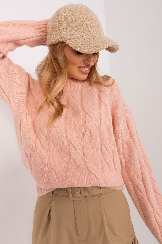 A woman wearing a dusty pink cable-knit sweater, a beige bouclé cap, and tan trousers, posing against a plain white wall.