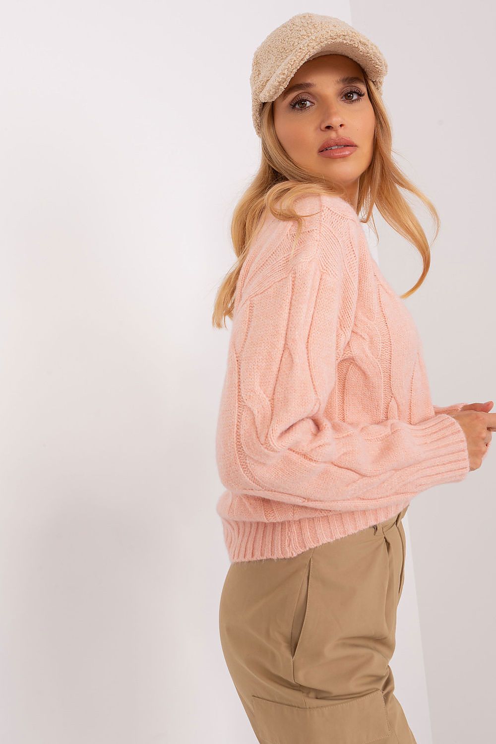 A woman wearing a light pink cable-knit sweater, beige pants, and a beige knit cap, photographed against a plain white studio background.