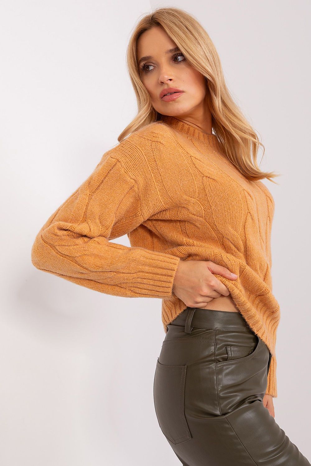 A woman wearing a cropped, mustard yellow cable-knit mock-neck sweater and dark olive green faux-leather pants, posing against a light gray studio background.