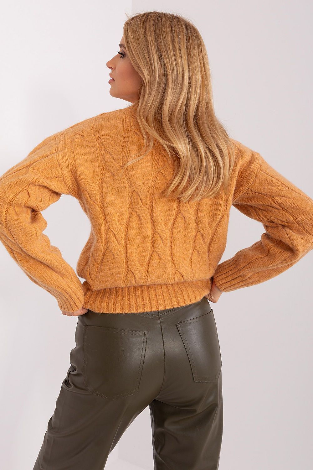A woman wearing a mustard yellow cable-knit sweater and dark olive green faux-leather pants, shown from the back against a plain light gray studio background.