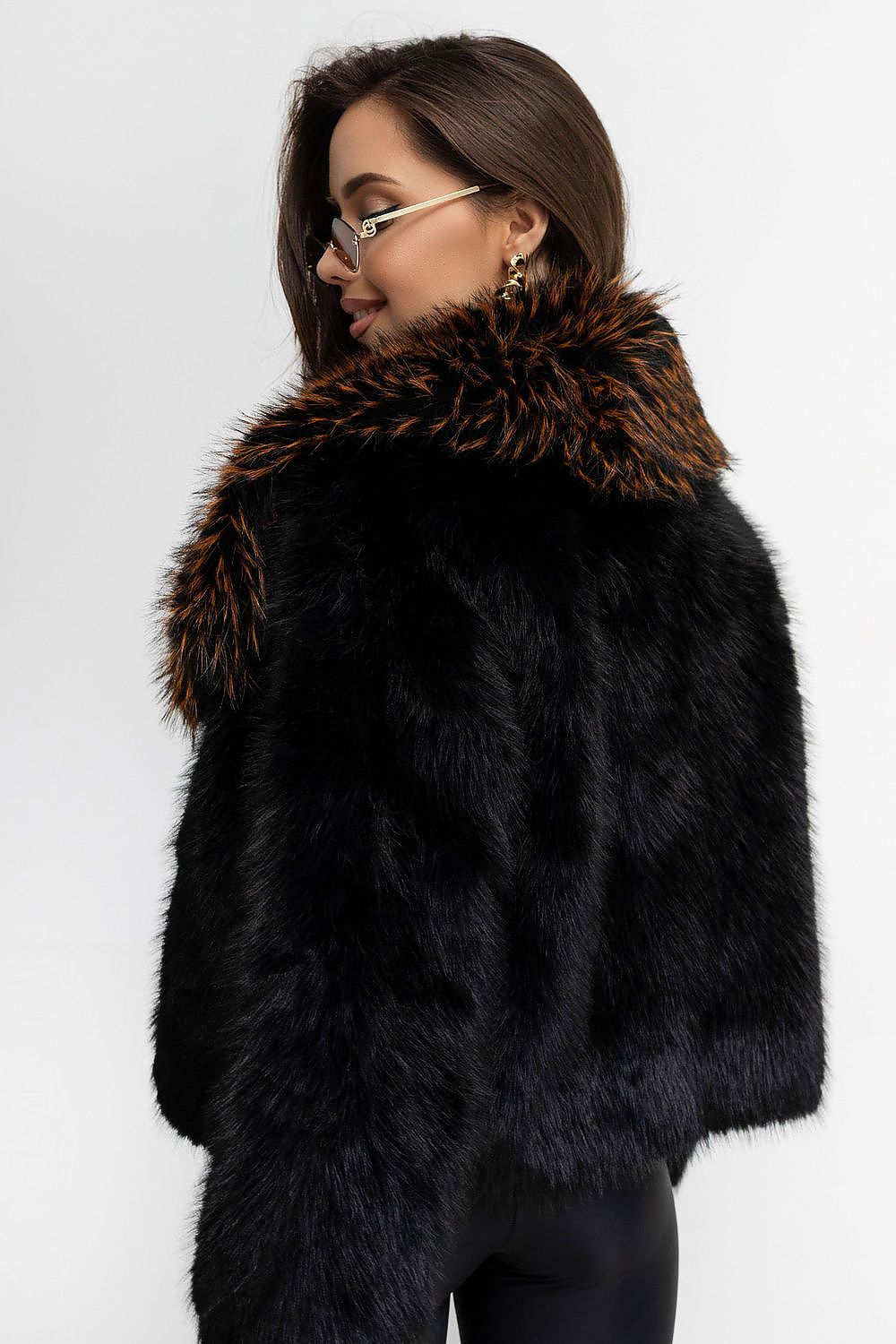 A woman wearing a black faux fur jacket with a large brown and black patterned fur collar, photographed against a plain light gray background.