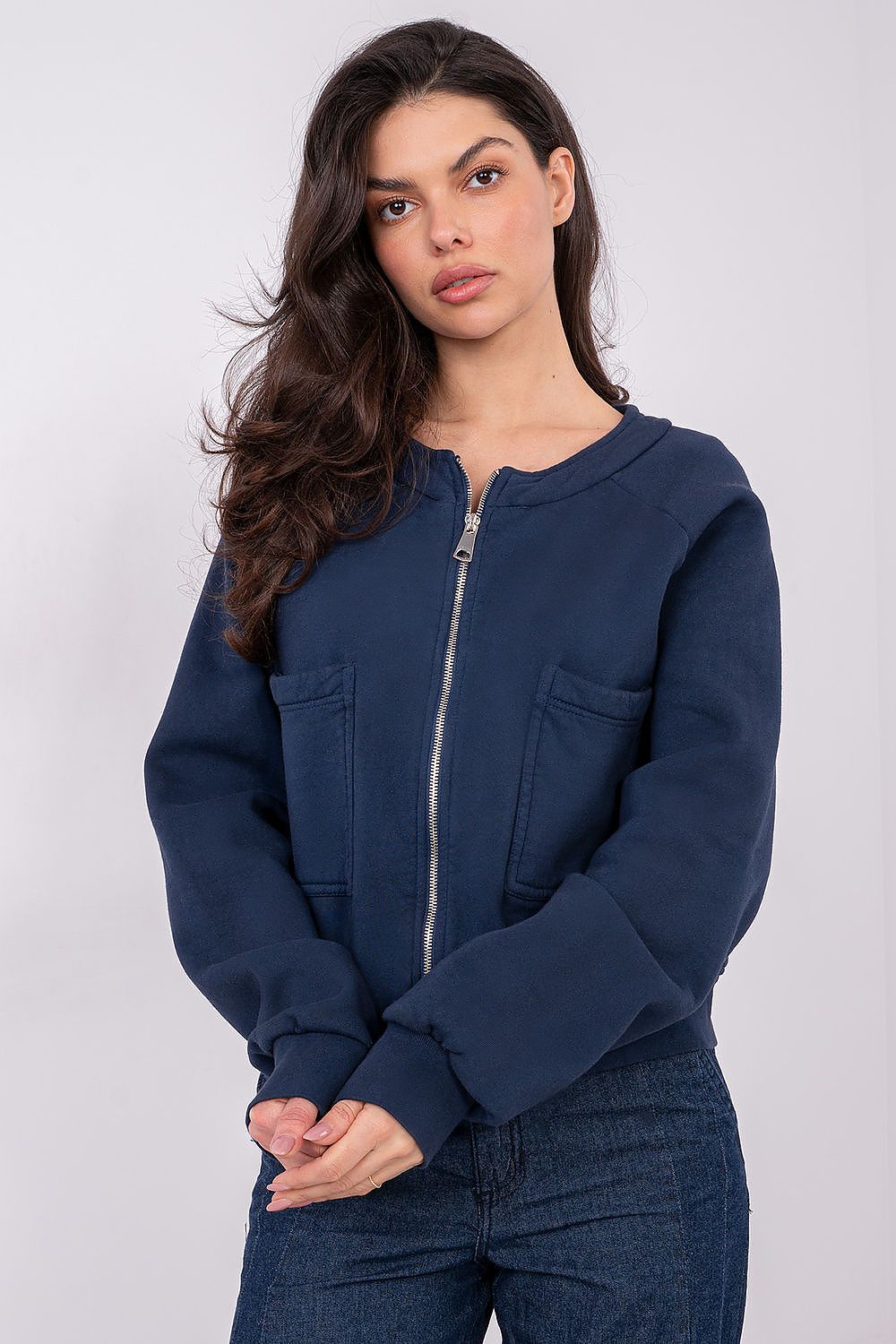 A woman modeling a navy blue cropped zip-up jacket with front pockets, paired with dark blue jeans, against a light gray studio background.