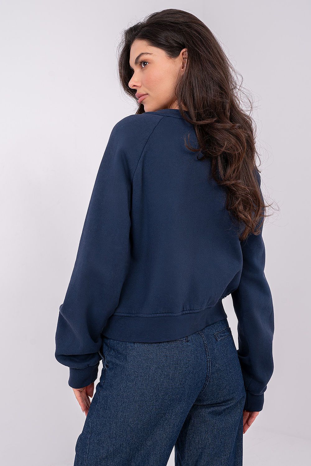 A woman wearing a navy blue cropped sweatshirt and blue jeans, photographed from a three-quarters back angle against a light gray studio background.