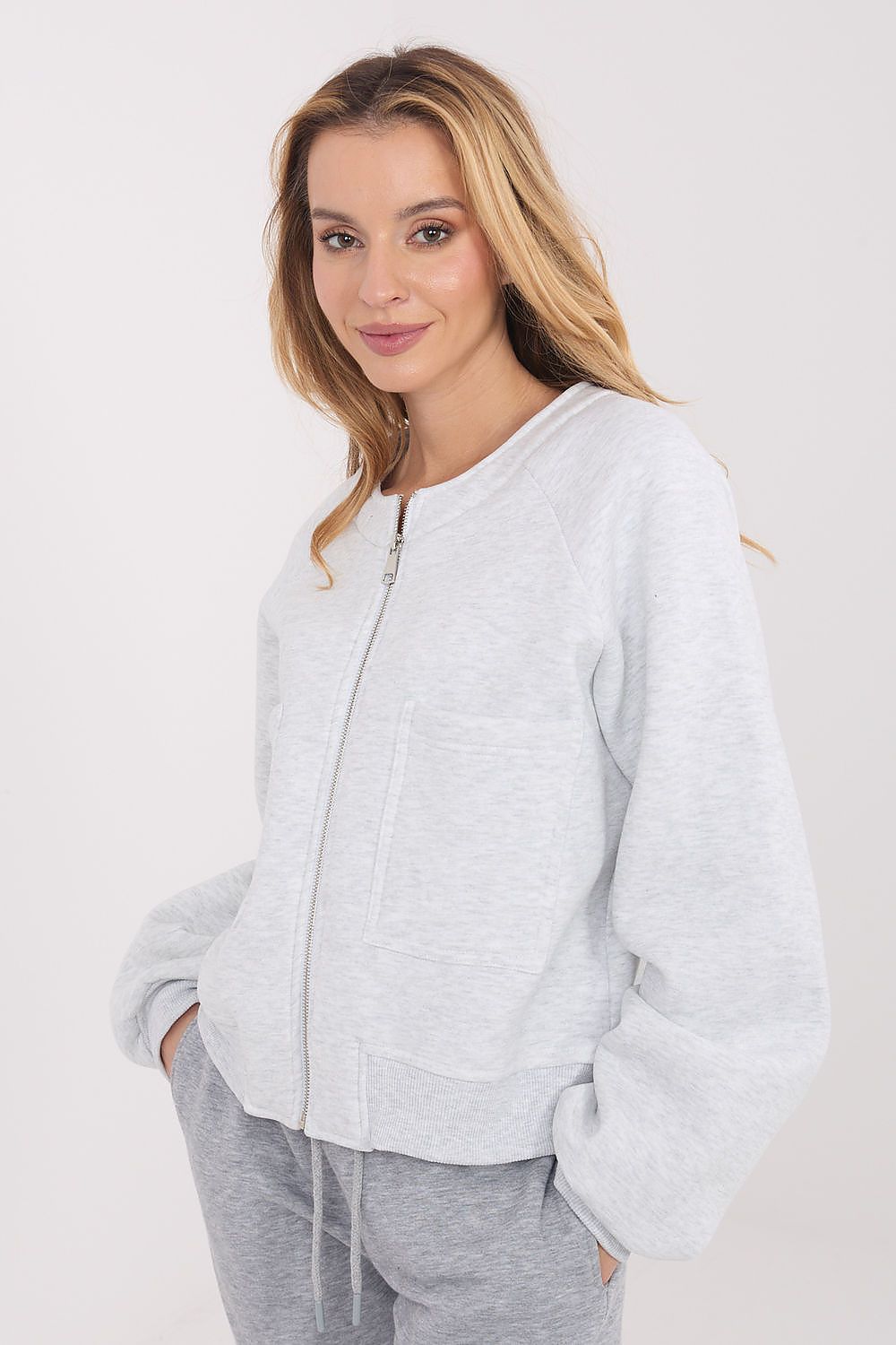 A woman wearing a light gray heather zip-up hoodie with a silver zipper and gray sweatpants, photographed against a plain light gray studio background.