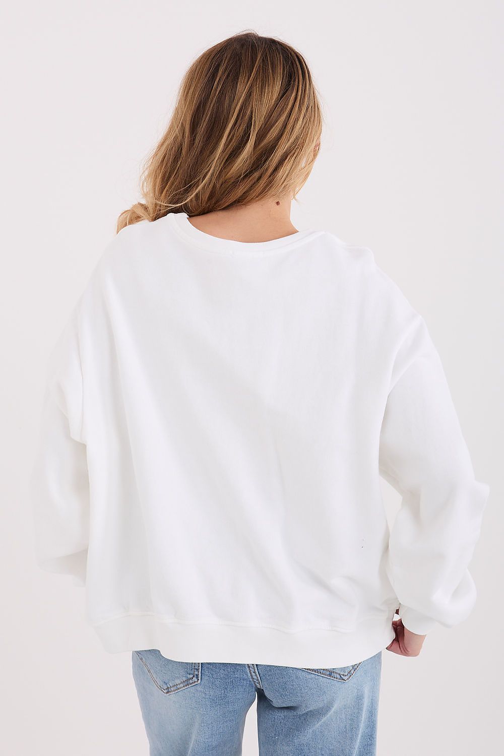 A woman viewed from behind wearing an oversized white crewneck sweatshirt and light blue jeans against a plain white background.