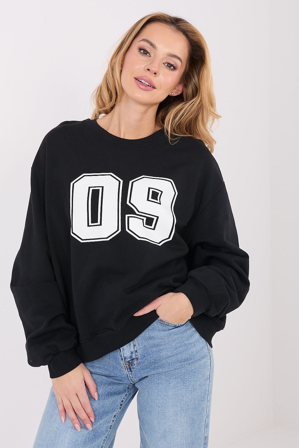 A woman wearing a black crewneck sweatshirt with large white '09' lettering outlined in black, paired with light blue jeans, against a plain light gray background.