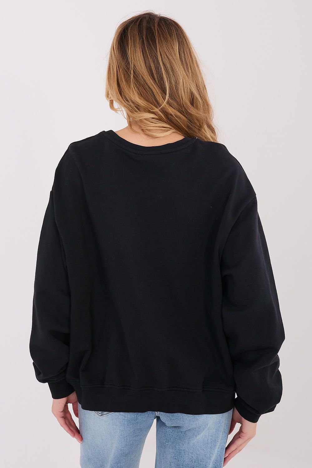 A woman viewed from the back wearing an oversized black crewneck sweatshirt and light blue jeans against a plain light gray studio background.