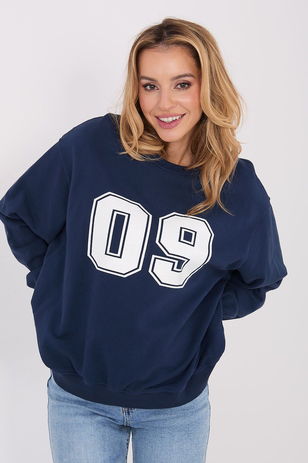 A woman wearing an oversized navy blue sweatshirt with a large white '09' graphic on the front, paired with light blue jeans, against a plain light gray background.