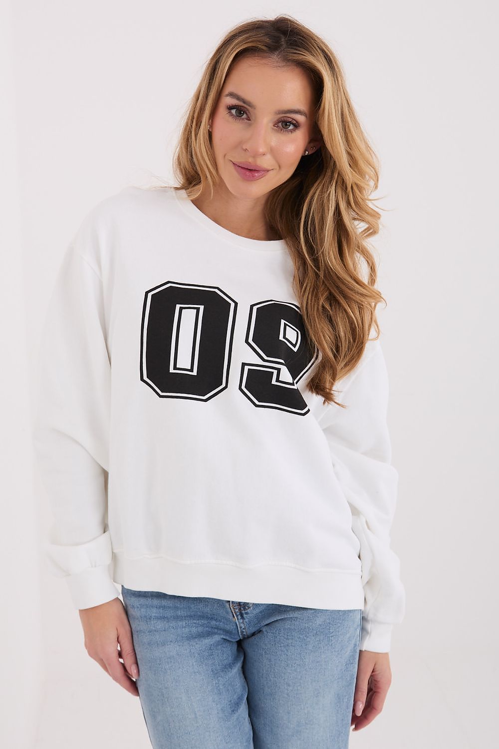 A woman wearing a white crewneck sweatshirt with a large black and white '09' graphic on the front, paired with light blue jeans, against a plain white studio background.