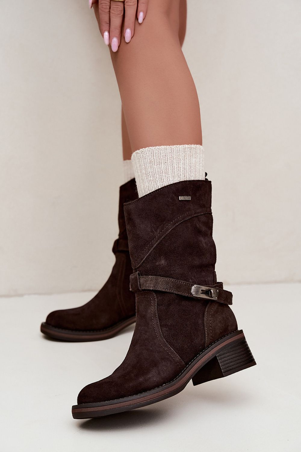 Dark brown suede ankle boots with a buckle strap detail and low block heel, shown on a model wearing white socks against a neutral beige background.