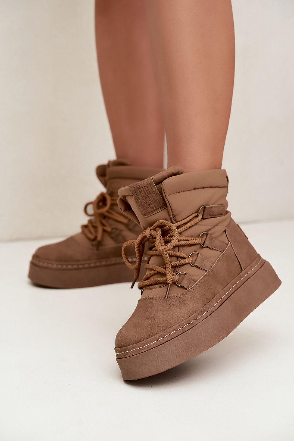 A pair of chunky platform ankle boots in a warm taupe-brown suede-like material, shown worn on a person's feet against a clean off-white studio background.