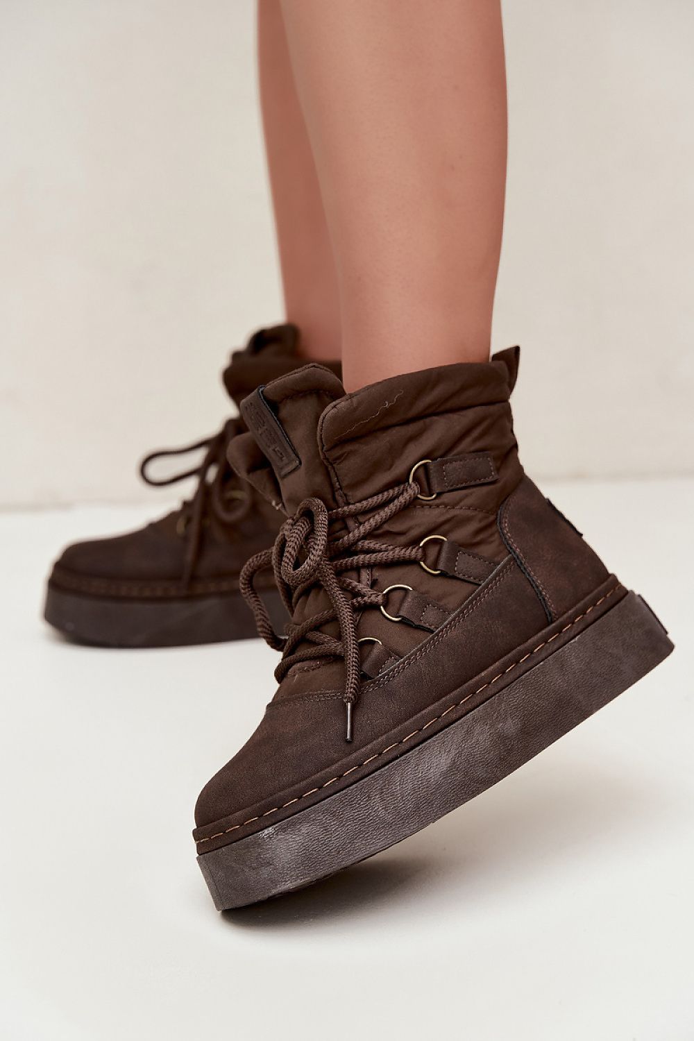 A pair of dark brown platform lace-up boots with metal eyelets, shown on a model's feet against a light beige studio background.