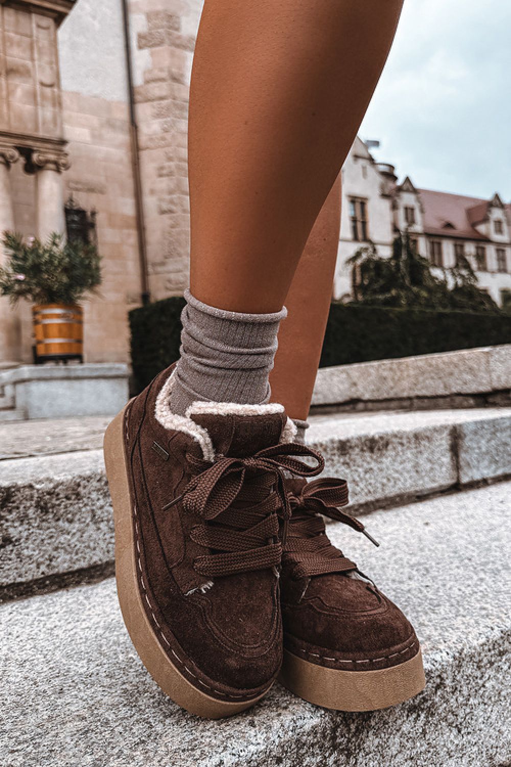 A person wearing brown suede low-top platform sneakers with a fuzzy white lining and gray socks, standing on stone steps outdoors with buildings in the background.