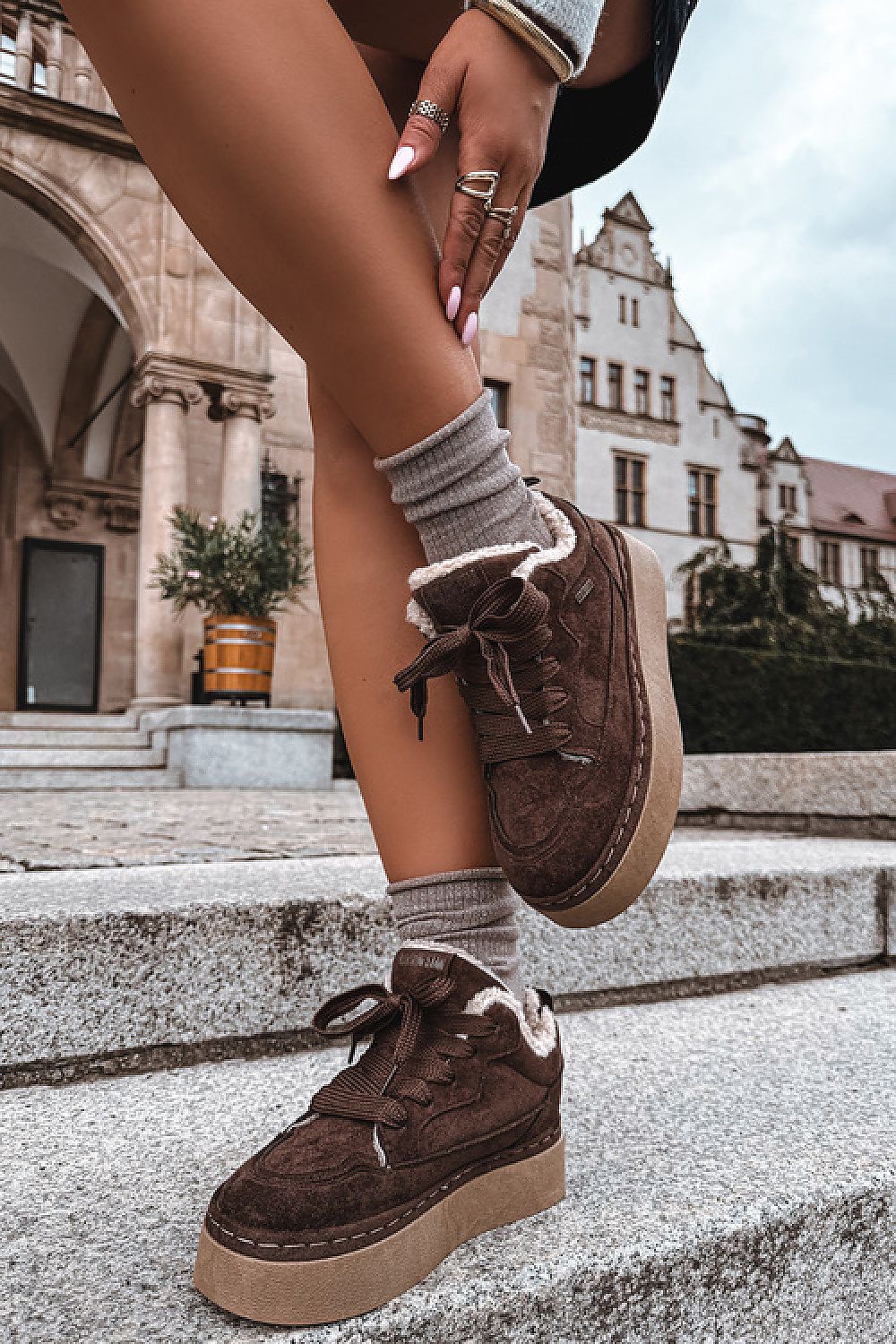 A person wearing brown suede low-top platform sneakers with white sherpa lining, styled with gray socks, standing on stone steps in front of historic buildings.