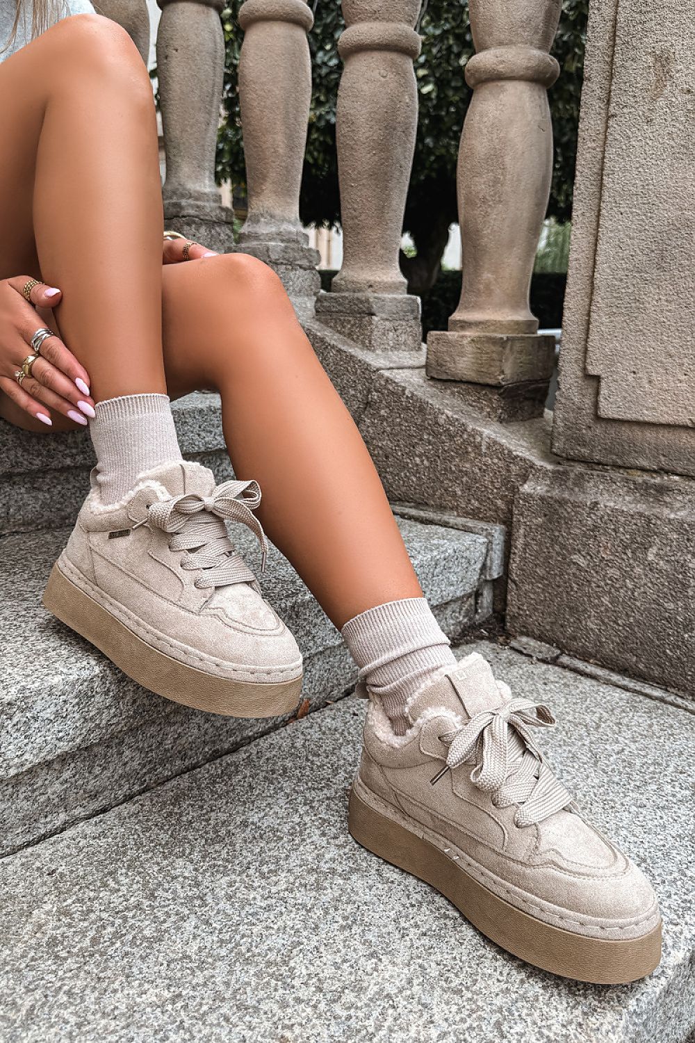 A person sitting on stone steps wearing chunky platform sneakers in a light beige or cream suede-like material with matching laces and ribbed socks.