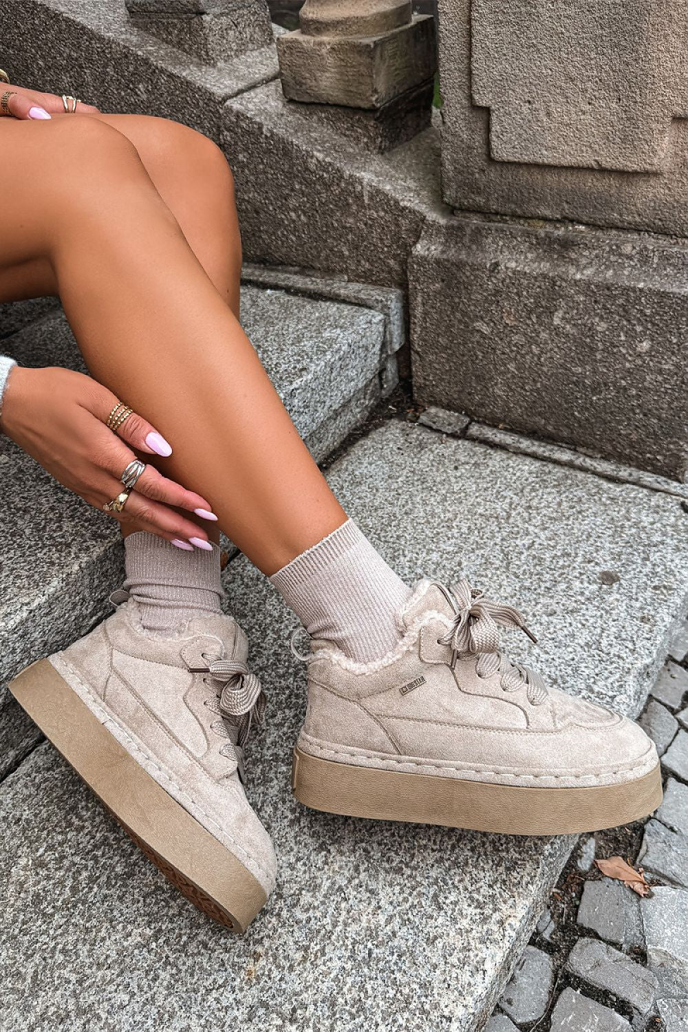 A person sitting on stone steps wearing beige platform sneakers with matching beige crew socks and multiple rings on their hand.