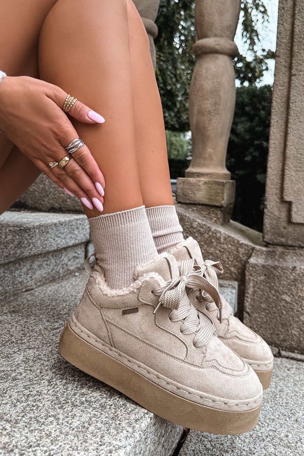 Beige low-top platform sneakers with fuzzy lining and matching lace-up socks, shown on a model sitting on stone steps outdoors.