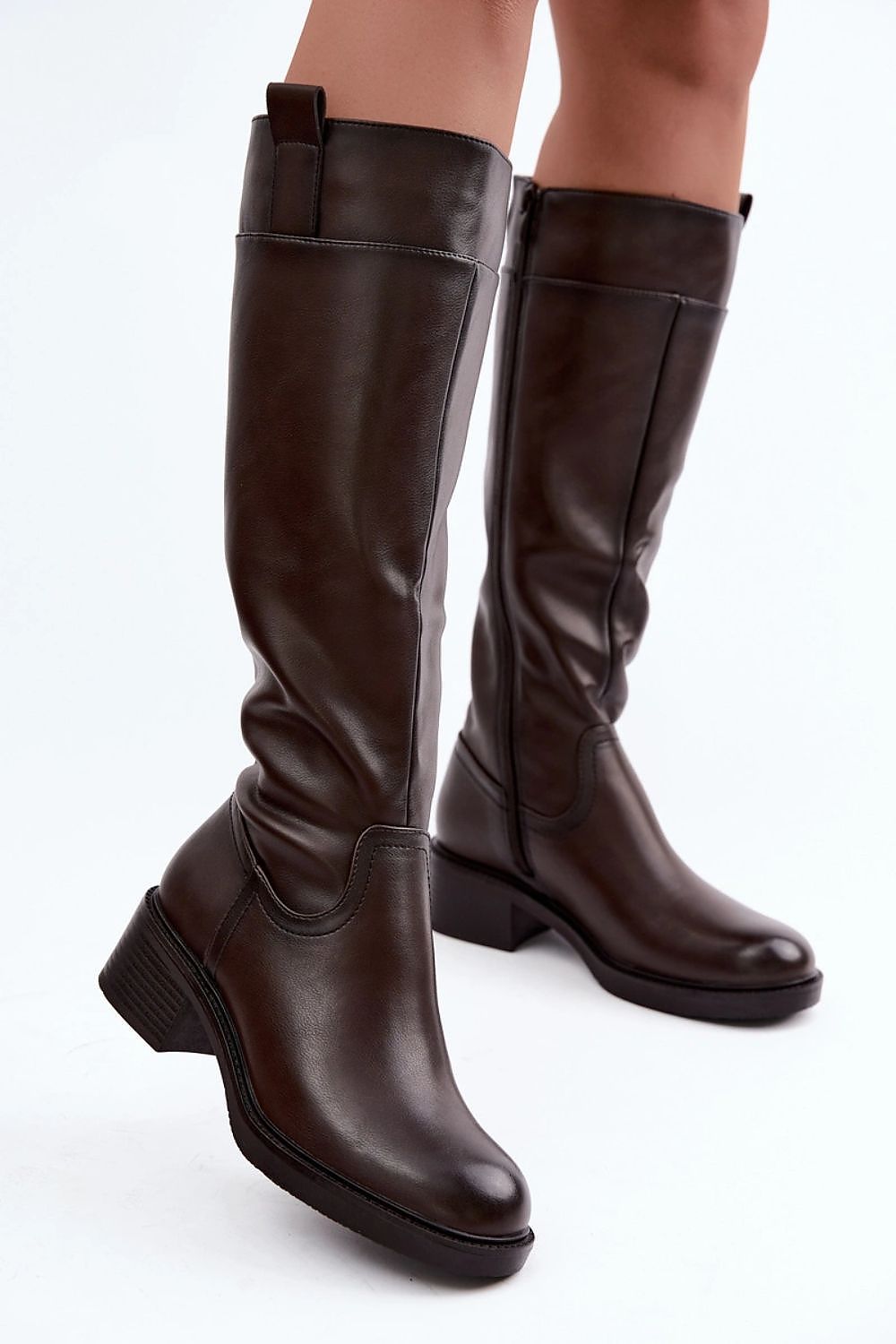 A pair of dark brown leather knee-high boots with low block heels, shown on a model's feet against a plain white studio background.