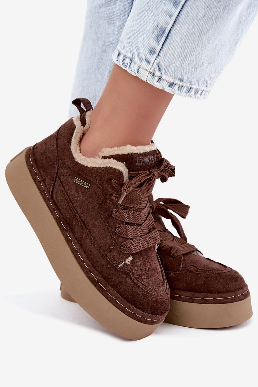 A pair of brown suede platform sneakers with a cream sherpa lining, shown on a model wearing light-wash blue jeans against a white background.