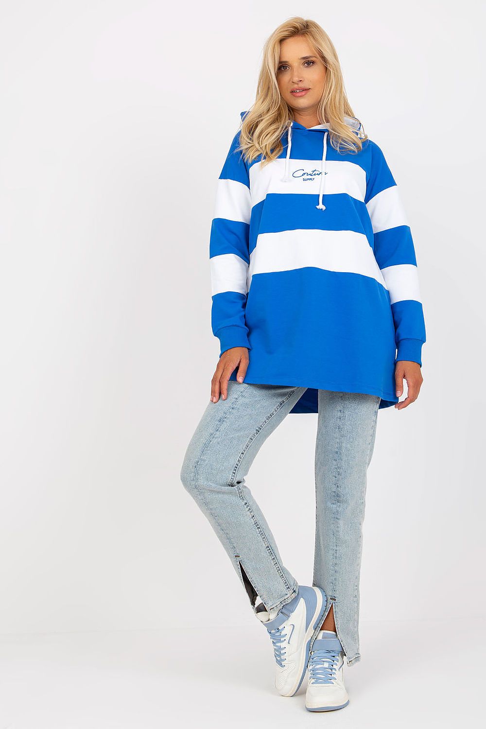 A woman modeling an oversized blue and white striped hoodie with a drawstring hood, paired with light-wash jeans and white sneakers, against a plain white studio background.