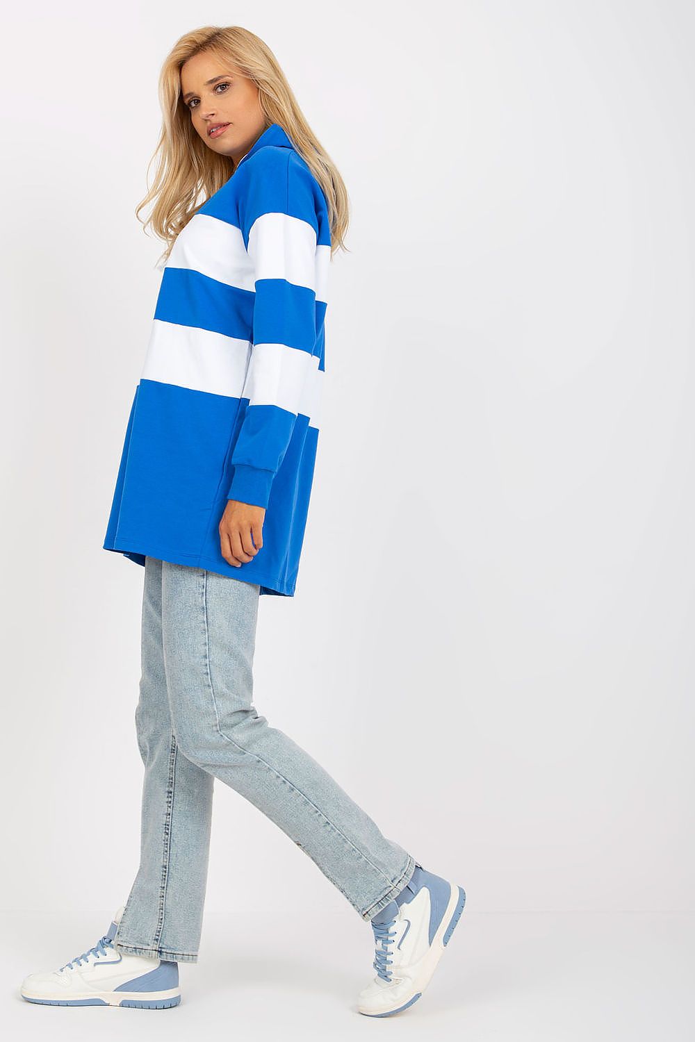 A woman modeling an oversized long-sleeve polo shirt with bold blue and white horizontal stripes, paired with light-wash jeans and white sneakers with blue accents, set against a plain light gray studio background.