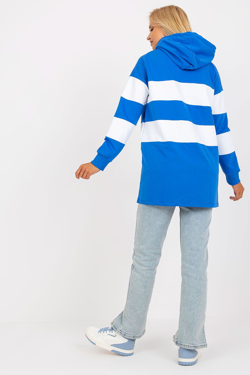 A woman shown from the back wearing a blue and white horizontally striped hooded sweatshirt, light-wash jeans, and white sneakers against a plain white studio background.