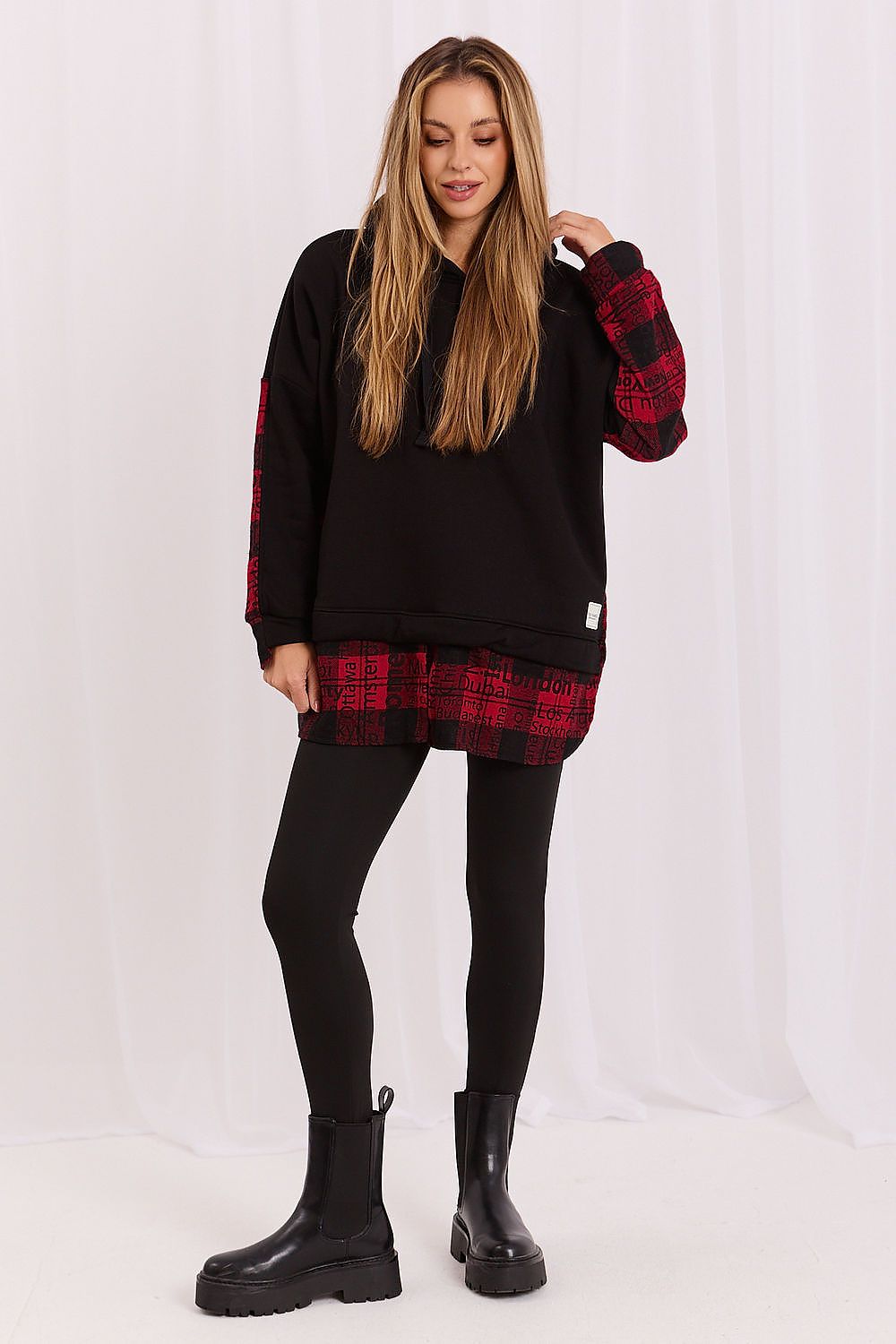 A woman modeling a black oversized sweatshirt with red and black plaid sleeves and hem, paired with black leggings and black boots, against a white studio backdrop.