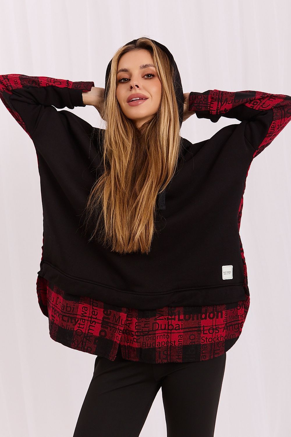 A woman modeling a black hoodie with red and black patterned sleeves, hem, and hood, set against a plain white background.
