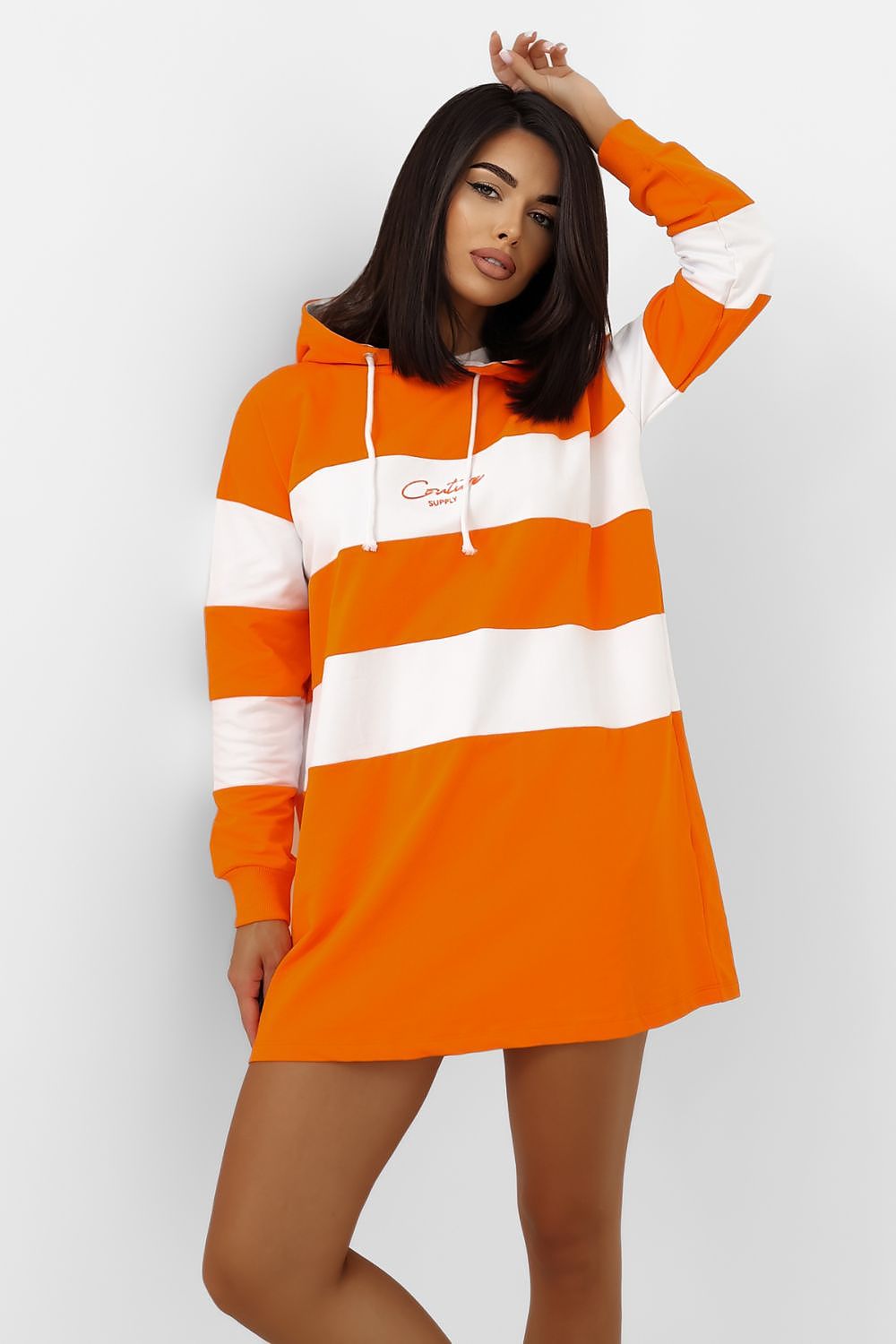 A woman modeling a bright orange and white striped hoodie dress with white drawstrings and a small script logo on the chest, set against a plain light gray studio background.