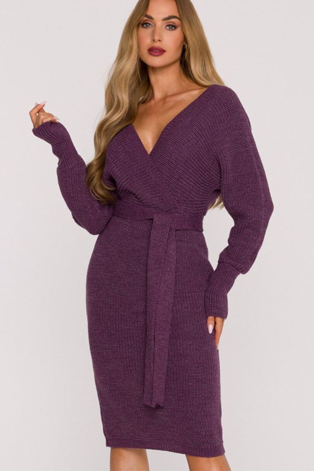 A woman modeling a ribbed knit long-sleeve bodycon dress in a deep plum purple color, featuring a wrap-style V-neckline with a matching tie belt at the waist.