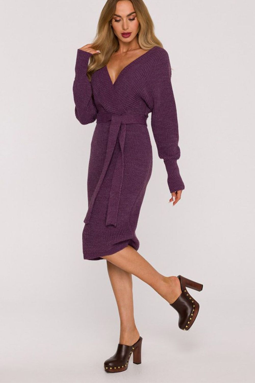A woman modeling a purple ribbed knit wrap dress with long sleeves and a tie waist, paired with dark brown clog heels, against a plain light gray studio background.
