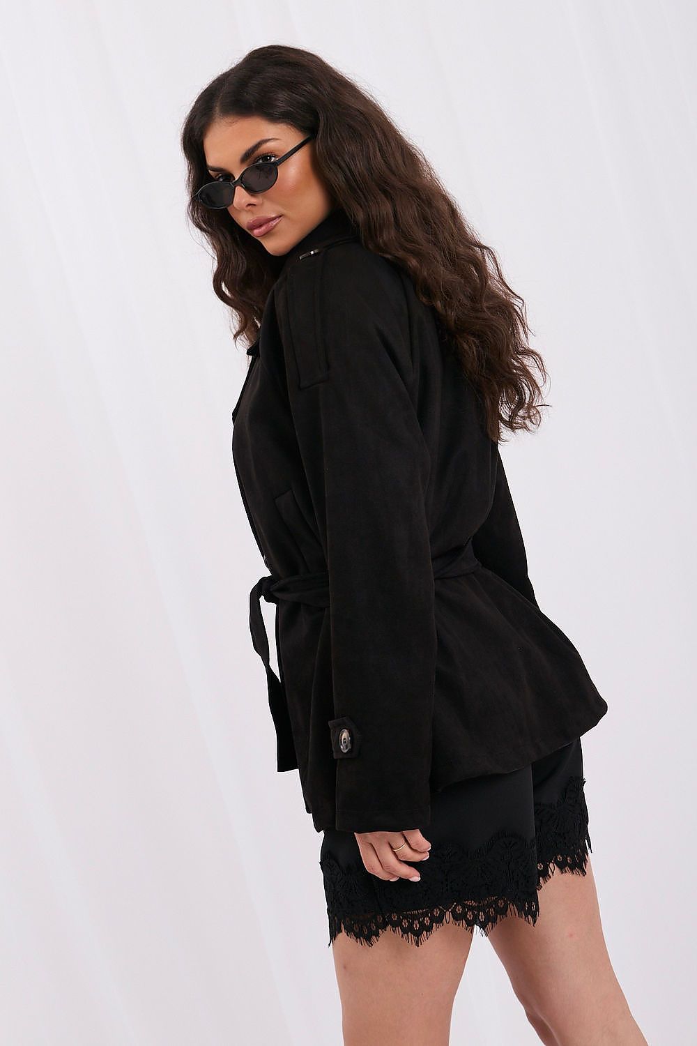 A woman wearing a black long-sleeve belted jacket and matching black lace-trim shorts, photographed against a plain white studio background.