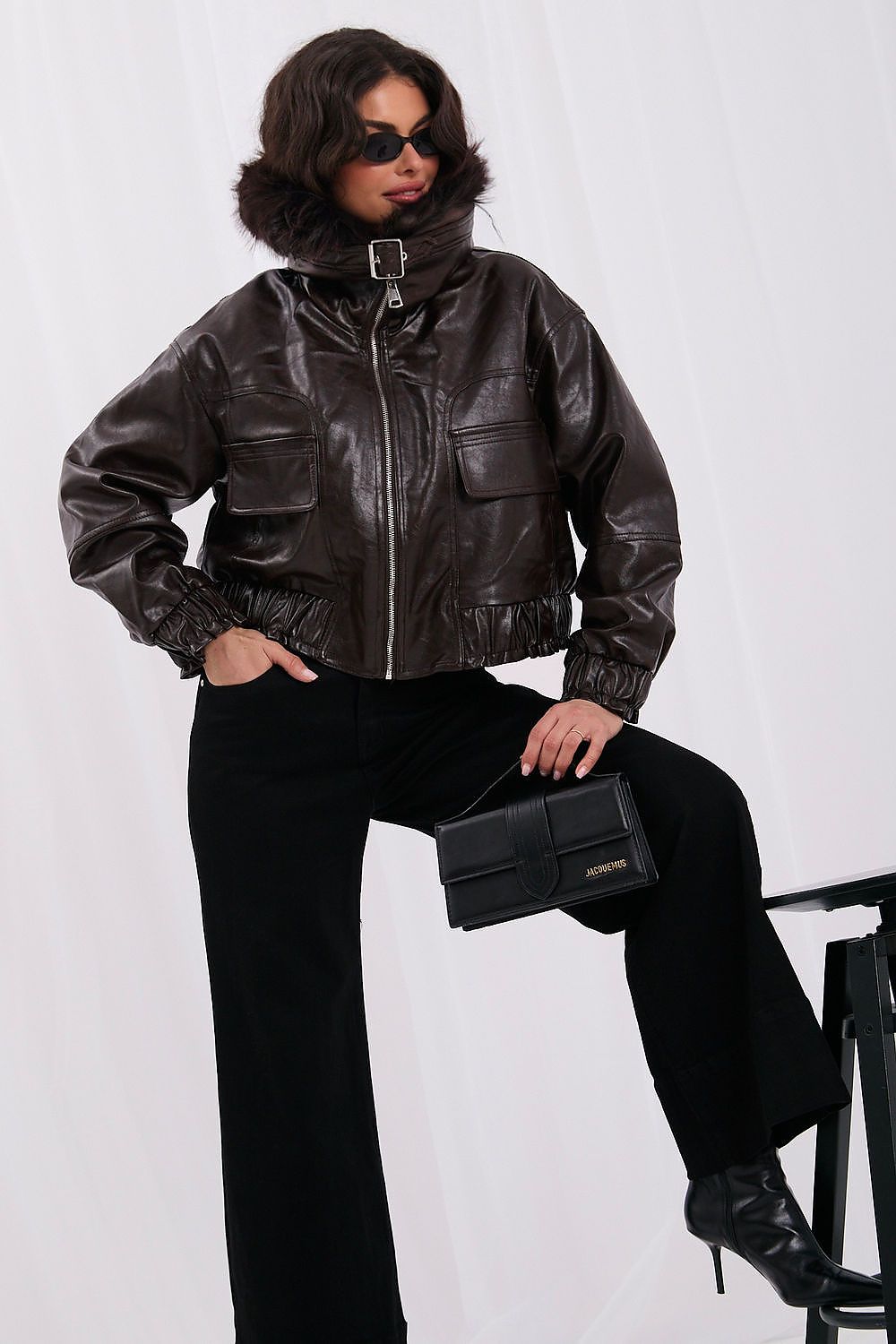 A model wearing a dark brown faux-leather moto jacket with a high collar, black wide-leg trousers, and black leather boots, holding a black clutch bag.