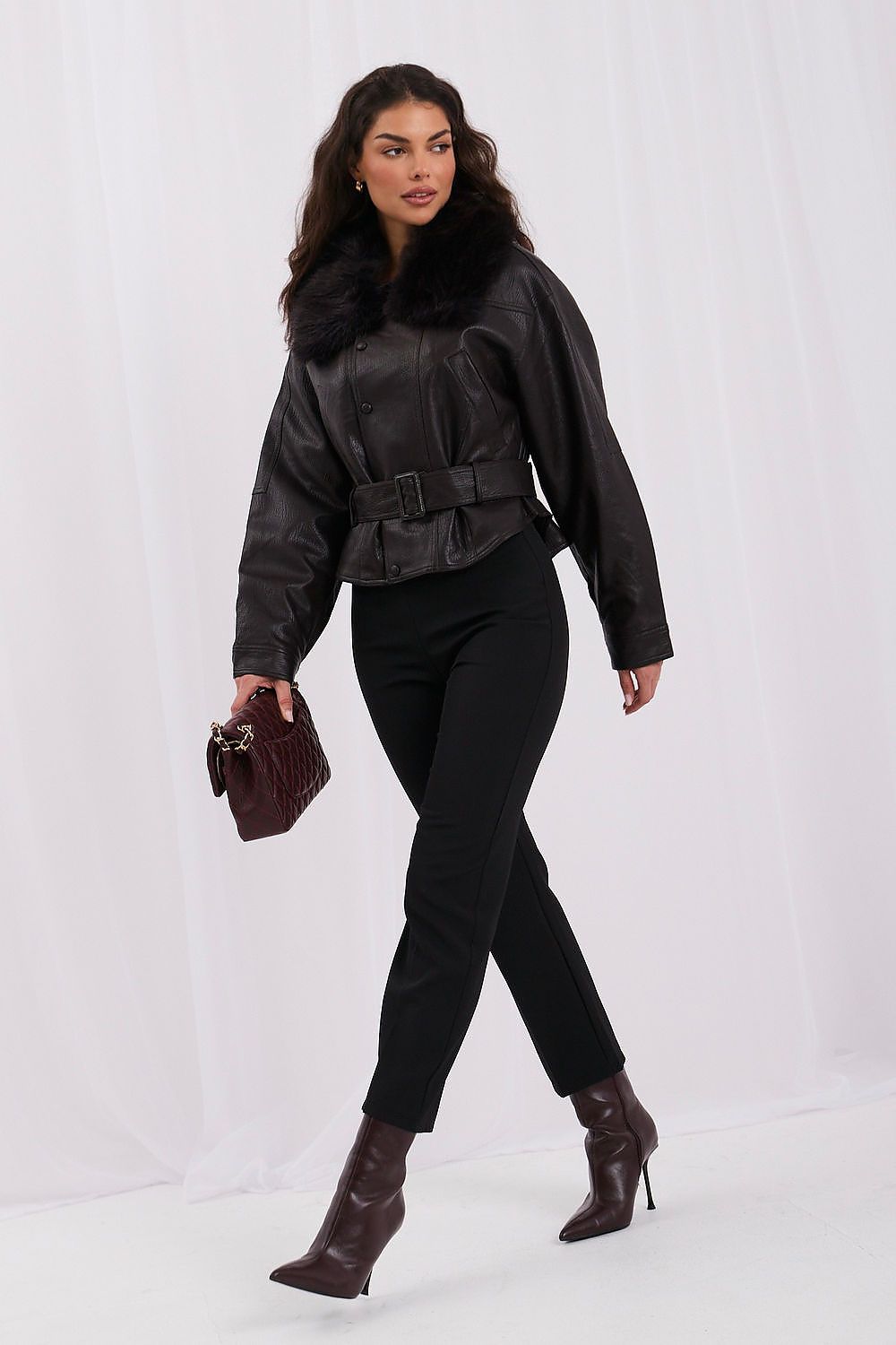 A woman models a black faux-leather jacket with a dark fur collar, belted at the waist, paired with black pants and dark brown heeled ankle boots, holding a small dark brown handbag against a white studio backdrop.
